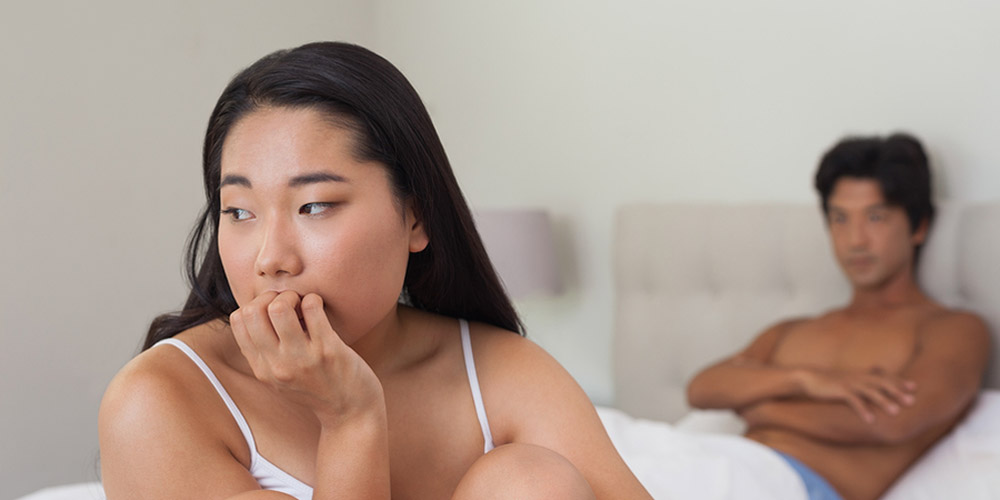 Boyfriend looking at upset girlfriend sitting on end of bed at h Boyfriend looking at upset girlfriend sitting on end of bed at home in bedroom